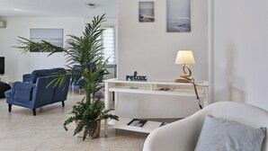 Ein modernes Wohnzimmer mit einer Couch, Sesseln, einem Couchtisch und einem Fernseher. Auf einem Konsolentisch befinden sich zwei Blumentöpfe und eine Lampe.
