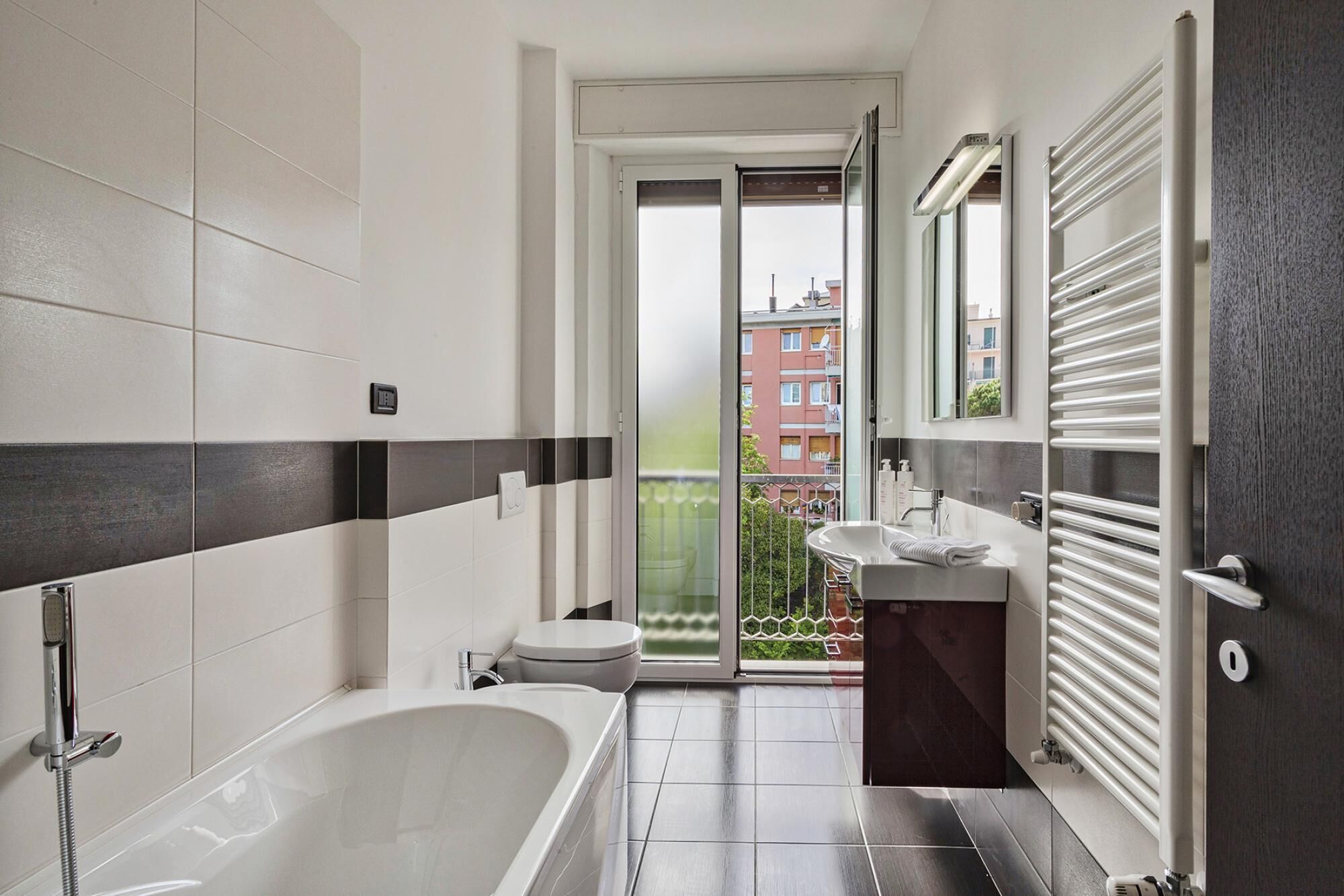 Een moderne badkamer met bad, toilet en wastafel, met uitzicht op een balkon en gebouwen.