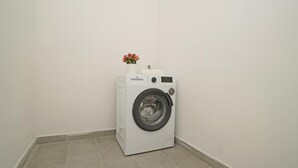 A compact, white front-loading washing machine in a corner of a room with plain white walls and tiled flooring. - San Carlo Borromeo 3 by Wonderful Italy (Soiano del Lago)