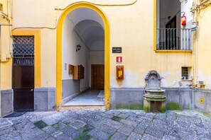 A narrow street with a yellow building featuring an archway entrance, a fire extinguisher, and a water fountain.