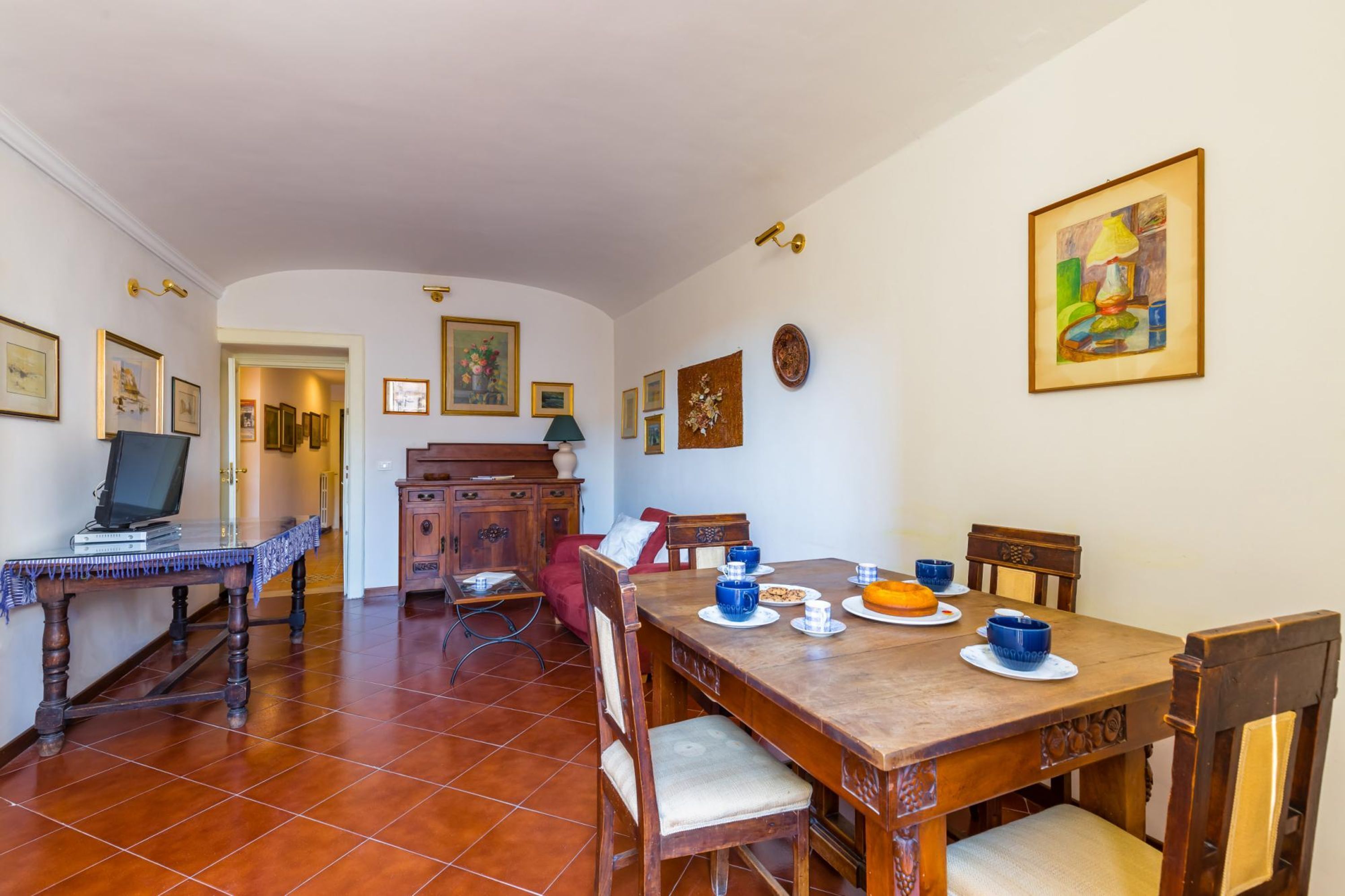 A dining area with a wooden table set for four, surrounded by chairs. There is a television on a stand to the left, and a wooden cabinet with drawers. The room has tiled flooring and white walls.