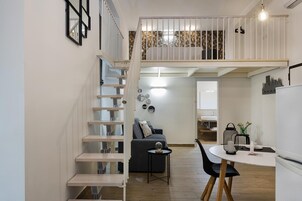A modern apartment with a staircase leading to a loft bedroom, a dining area with a round table, and a kitchen visible in the background.