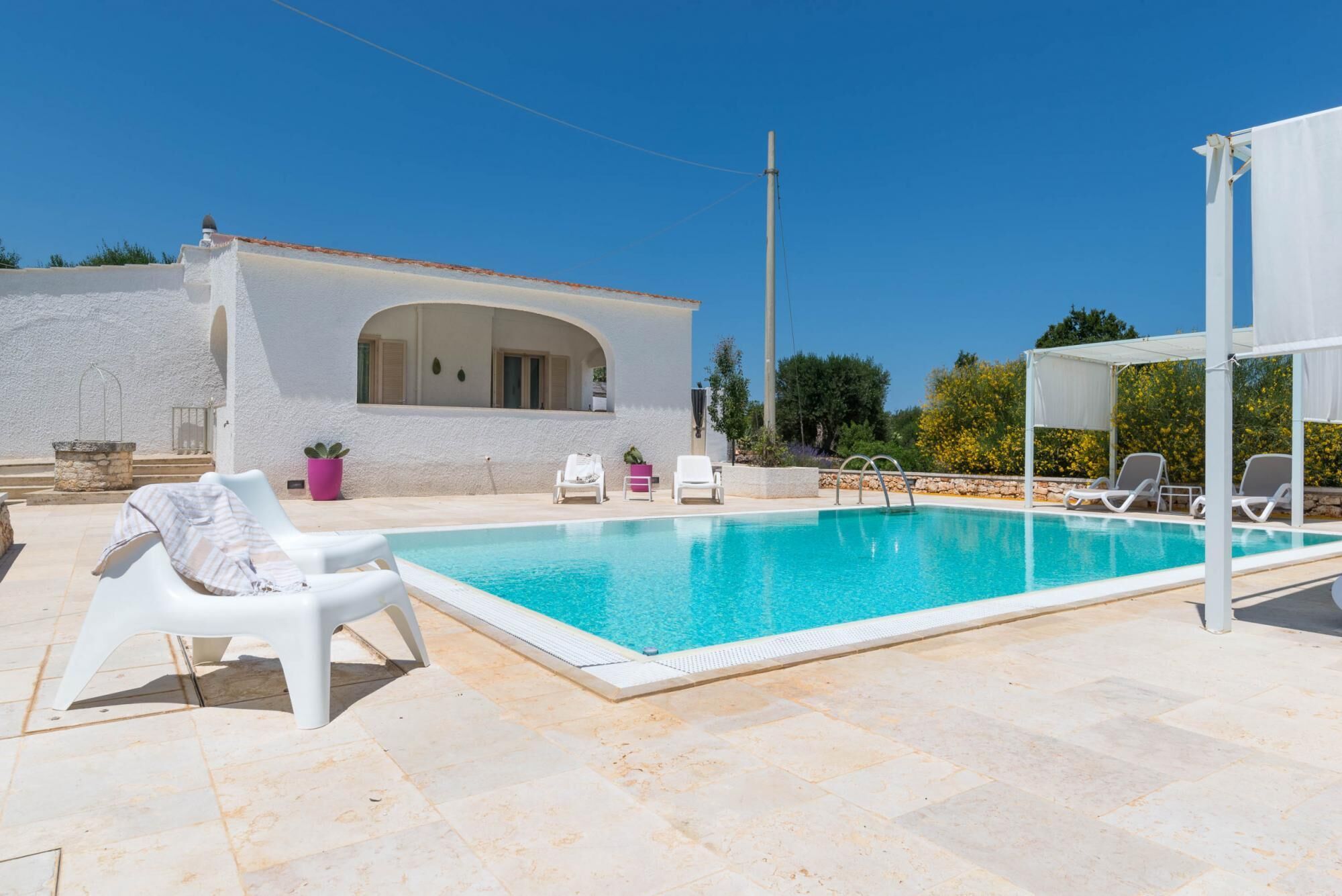 Una piscina con lettini e zona riparata, circondata da un edificio bianco con una finestra ad arco e un pozzo in pietra.