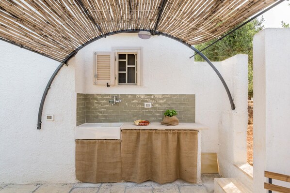 Un petit espace de cuisine extérieur, à l’abri, comprenant un évier, une fenêtre avec volets et une structure en bambou au-dessus.