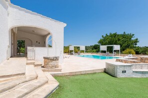 Un espace piscine agrémenté d’une structure en pierre, d’un espace salon couvert et d’un bâtiment blanc doté d’une entrée en arc de cercle.