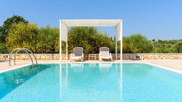A clear swimming pool with two lounge chairs under a pergola, surrounded by stone walls and greenery. - Villa d Itria Trullo Pool by Wonderful Italy (Ostuni)