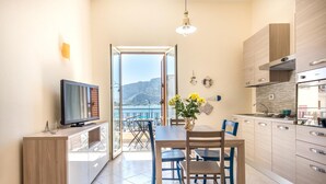 A modern kitchen with a view of the sea, a dining area with blue chairs, and a TV on a wooden cabinet.