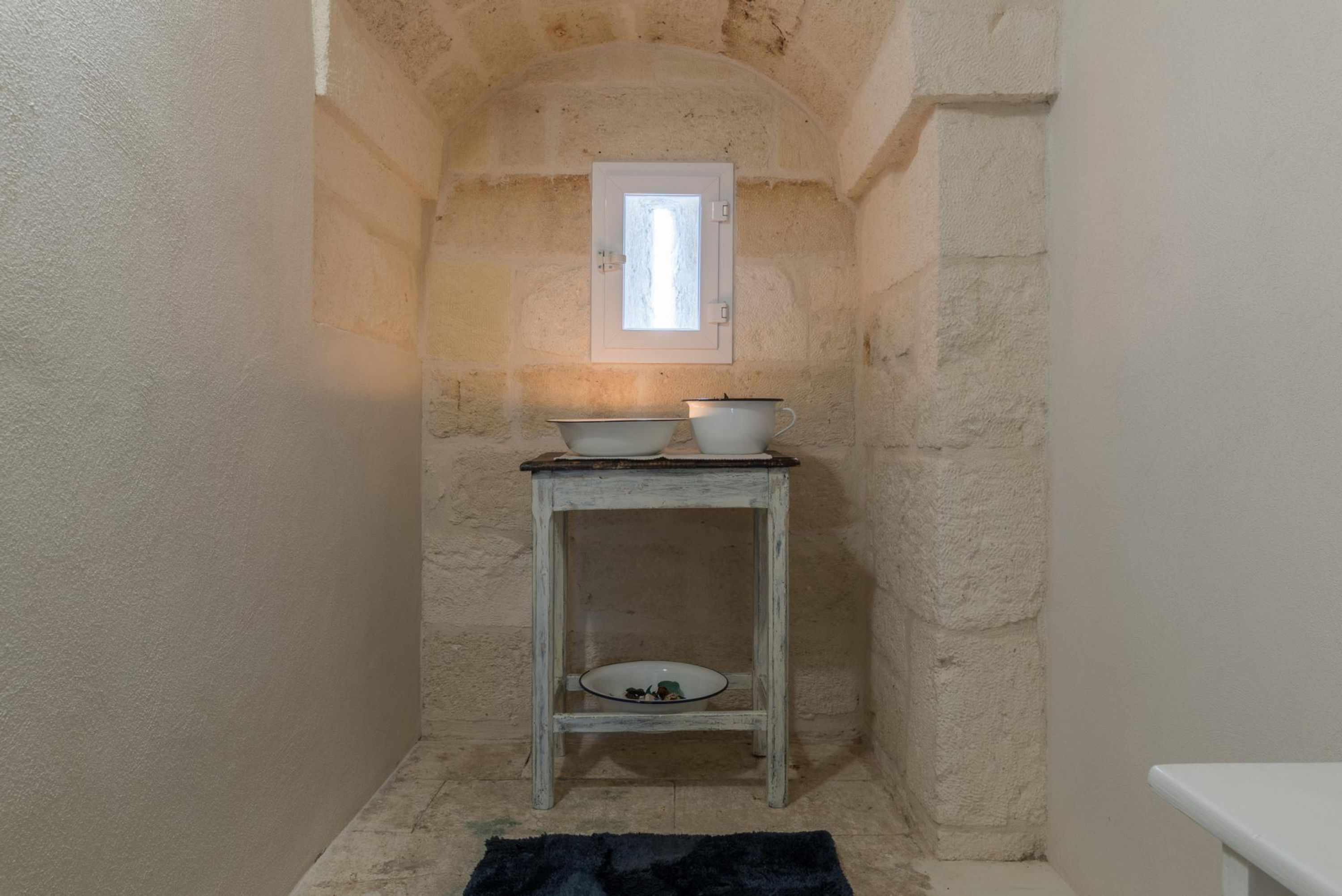 Une petite salle de bain aux murs de pierre, avec une petite fenêtre, une table en bois rustique sur laquelle sont posés des bols, et un tapis noir sur le sol.