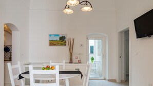 A dining area with a white table, chairs, and a fruit bowl. There is a painting on the wall and a television mounted on the wall. - Casetta Biancapietra by Wonderful Italy (Ostuni)