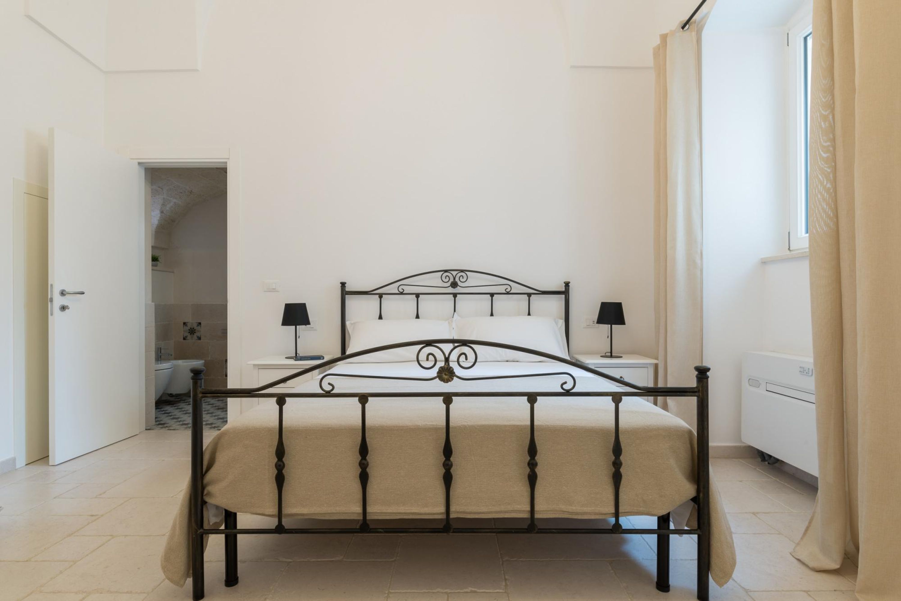 A bedroom with a metal bed frame, a bedside table with a lamp, and a window with curtains.