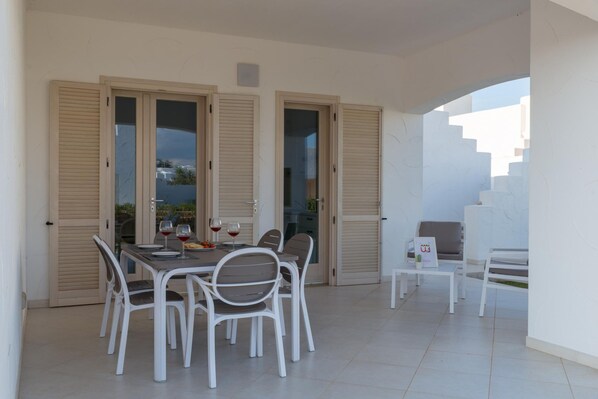Un coin repas avec une table dressée pour quatre personnes, entourée de chaises blanches. On y trouve deux portes avec volets, une petite table sur laquelle est posé un vase, et un canapé.