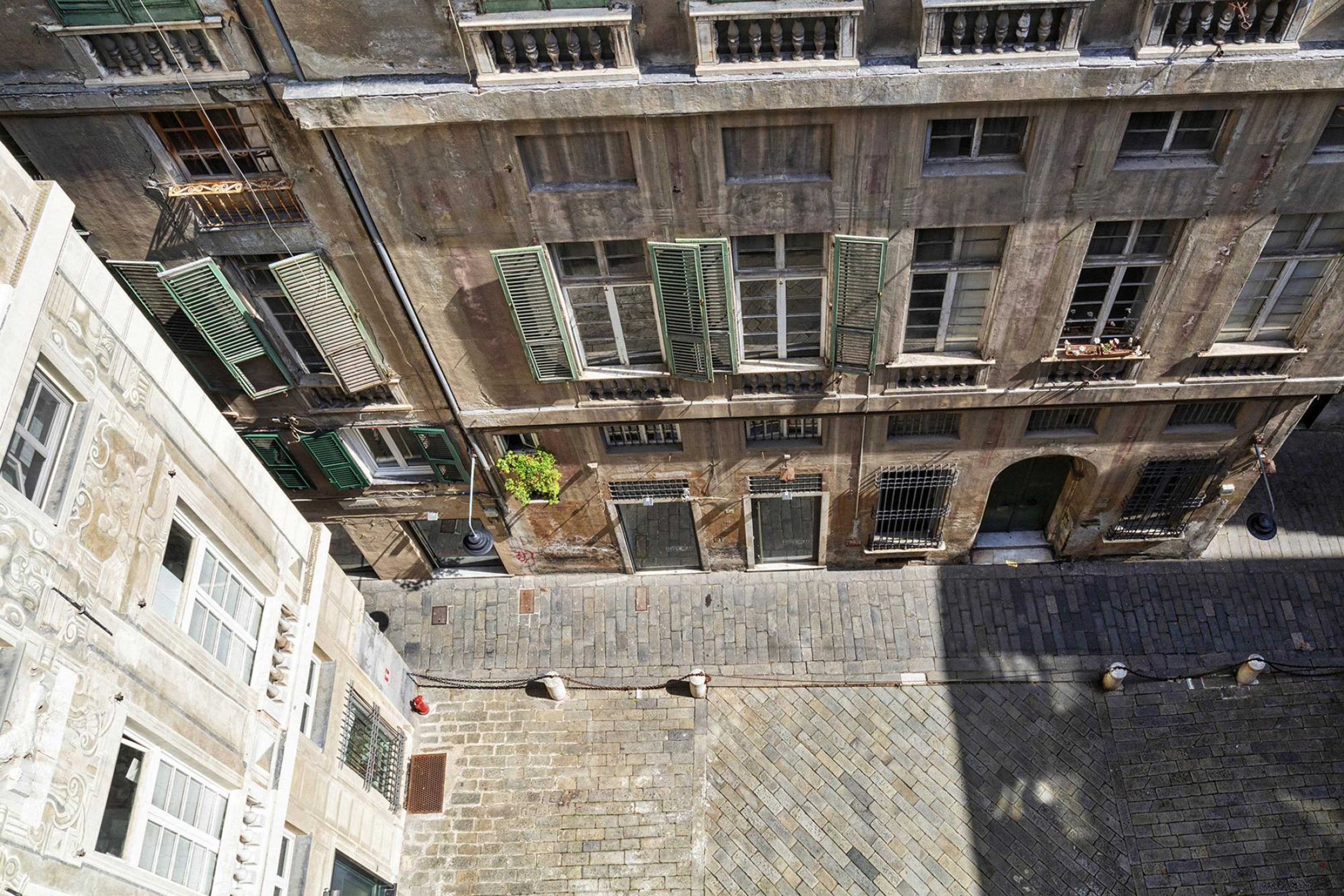 A multi-story building with a brick facade, multiple balconies with green shutters, and a paved ground floor.