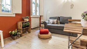 A cozy living room with a grey sofa, a wooden coffee table, and a red cushion. There are potted plants on a wooden shelf and a floor lamp. - Corte Galluzzi in Basilica by Wonderful Italy (Bologna)