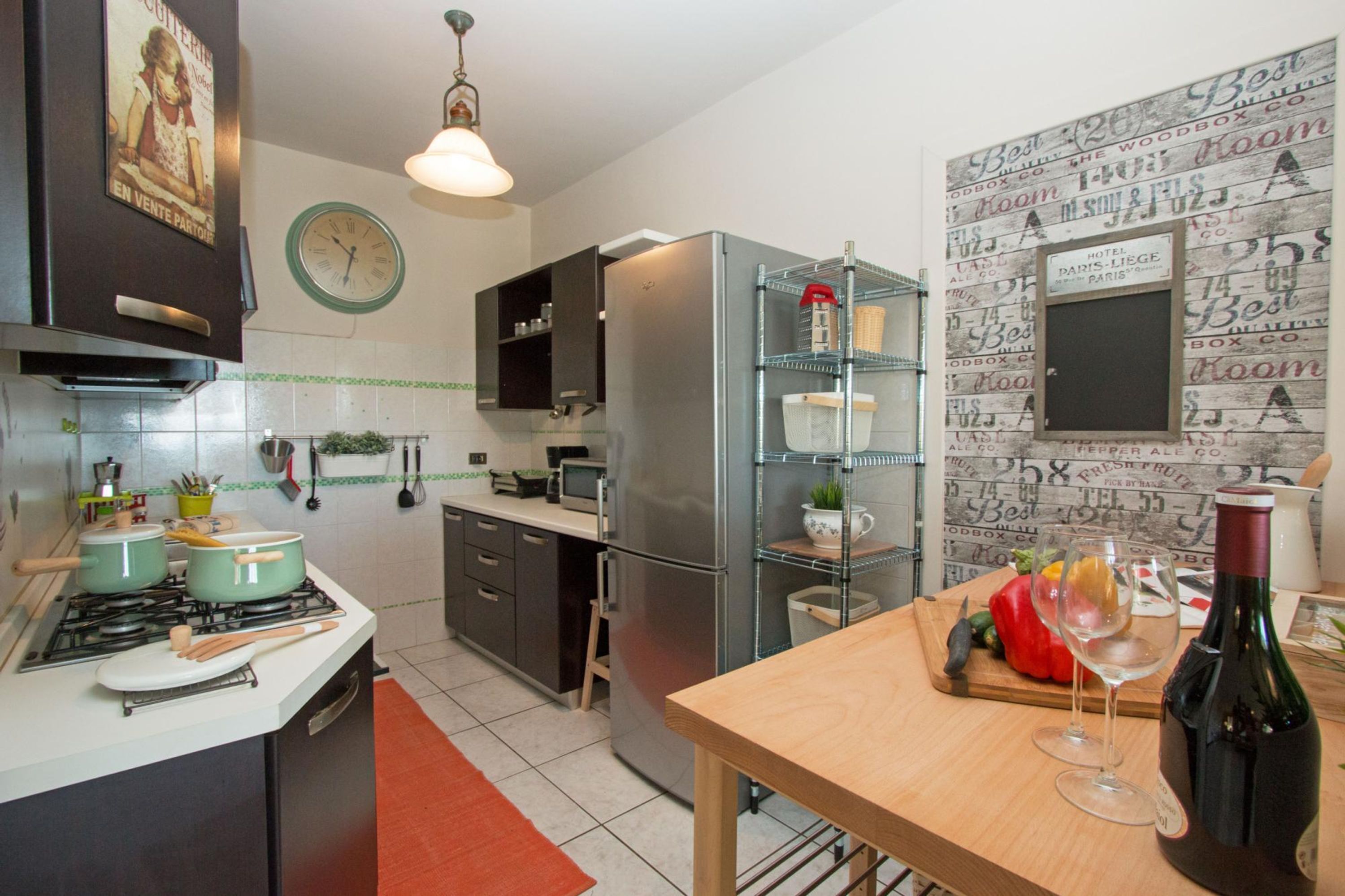 A modern kitchen with a refrigerator, stove, and microwave. There is a wooden table with a wine bottle and glasses.