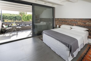 A bedroom with a large bed, a stone headboard, a sliding glass door to a balcony, and a view of greenery outside.