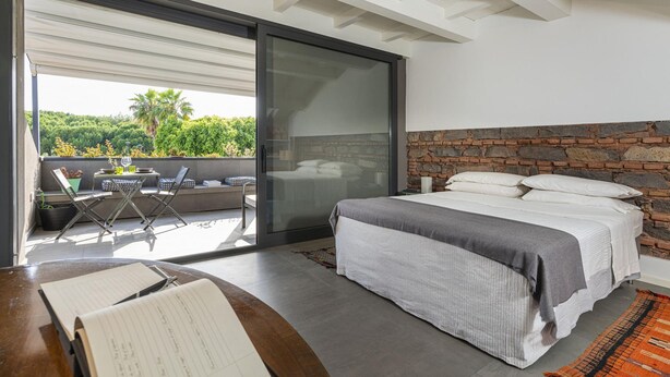Een slaapkamer met een groot bed, een accentmuur van steen en een schuifdeur naar een terras met uitzicht op groen.
