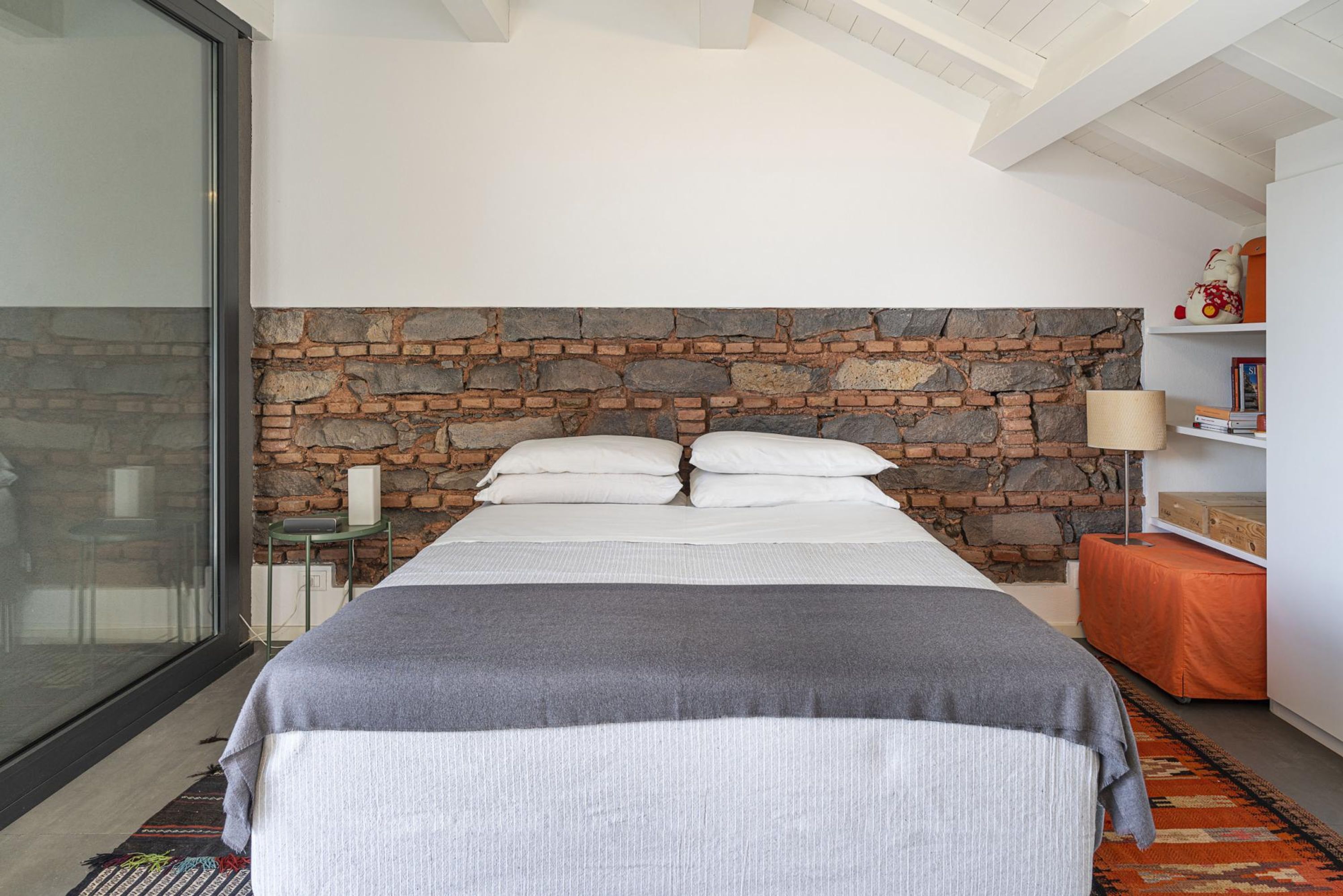 Une chambre à coucher avec une tête de lit en pierre, un lit recouvert de draps blancs, une table de chevet avec une lampe et une bibliothèque.