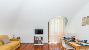 Ein gemütliches Wohnzimmer mit gelbem Sofa, einer Bücherwand, einem Flachbildfernseher und einem Essbereich mit Tisch für zwei Personen.