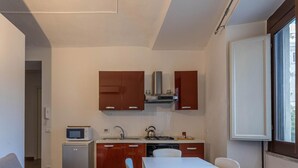 A compact kitchen with a microwave, stove, and sink. There is a small refrigerator and wooden cabinets. A white table and chair are positioned near the window. - Casetta Borsa by Wonderful Italy (Palermo)