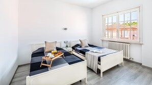 A small, bright room with two beds, a window with a view of buildings, and a radiator. - Il Quadrifoglio D4 con Piscina by Wonderful Italy (Sirmione)