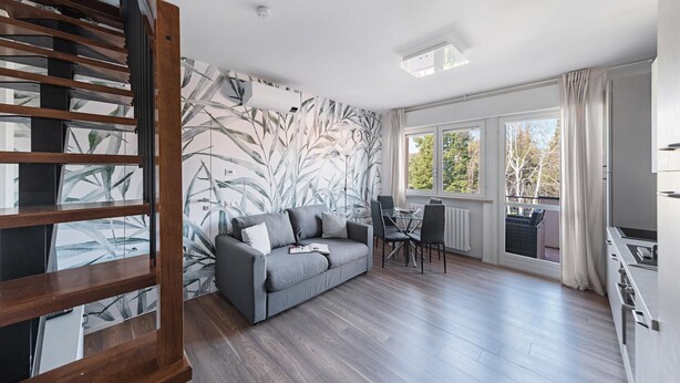 A modern living room with a grey sofa, a dining table with chairs, a staircase with wooden railings, and large windows with curtains. - Maisonnette Il Todeschino B12 by Wonderful Italy (Sirmione)