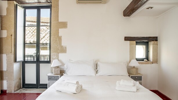 A hotel room with a large bed, two bedside tables with lamps, a window with a view of buildings, and a small mirror on the wall. - Le Terrazze Dell Olivo by Wonderful Italy (Syracuse)