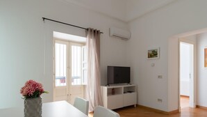 Un salon moderne avec un téléviseur à écran plat posé sur un meuble blanc, une table à manger avec des chaises, une grande fenêtre avec des rideaux et une porte donnant sur une autre pièce.
