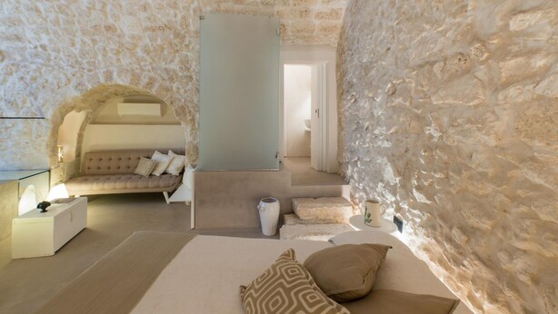 A cozy bedroom with a stone wall, a comfortable sofa, a bed with pillows, and a nightstand with a lamp.