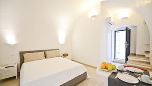 A bedroom with a large bed, a bedside table, a small table with fruits, and a view of a staircase. - Scirocco Apartment With Terrace by Wonderful Italy (Ostuni)
