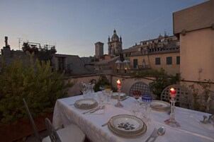 Montaje de comedor al aire libre con una mesa dispuesta para dos personas, rodeada de follaje y vistas a un paisaje urbano con edificios históricos.