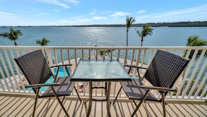 Un balcon avec une table en verre et deux fauteuils en osier, donnant sur une étendue d’eau et des palmiers.