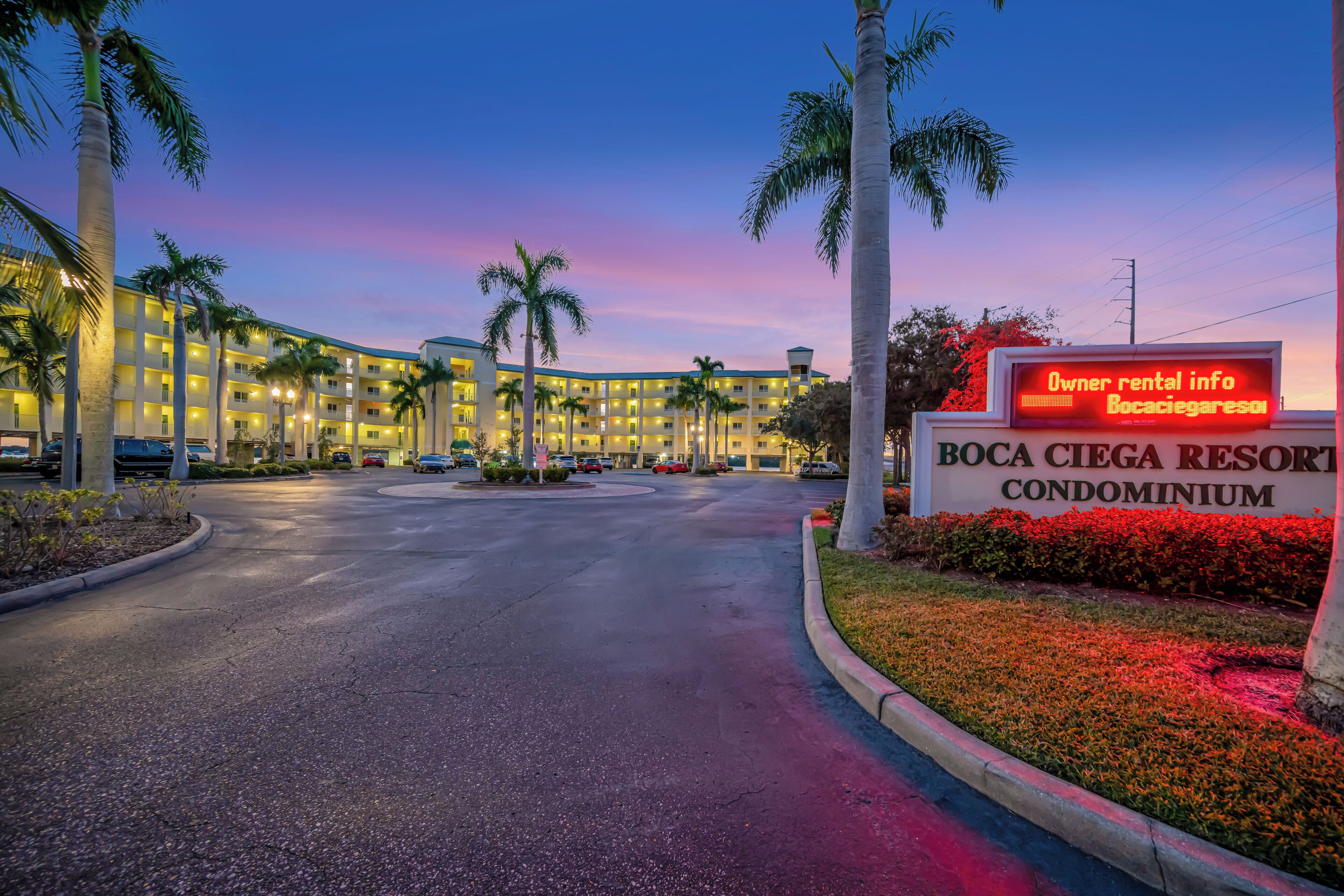 A hotel entrance with a sign for Boca Ciega Resort Condominium, featuring a large building, palm trees, and a well-lit driveway.