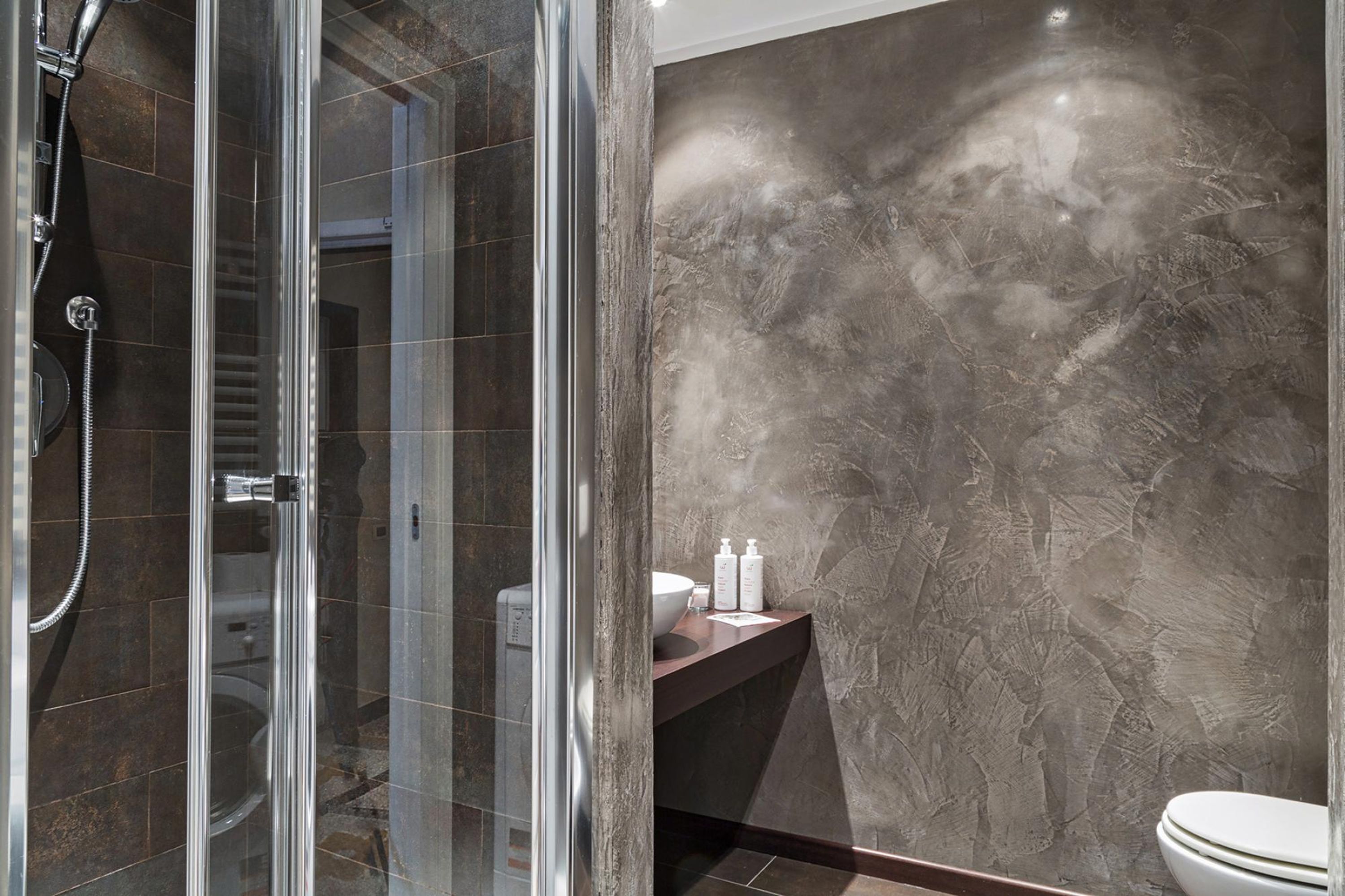 A modern bathroom with a glass-enclosed shower, a marble wall, and a dark countertop with toiletries.