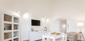 A modern living room with a white sofa, a dining table with chairs, a TV mounted on the wall, and a floor lamp. - Villino a Fontane Bianche by Wonderful Italy (Syracuse)