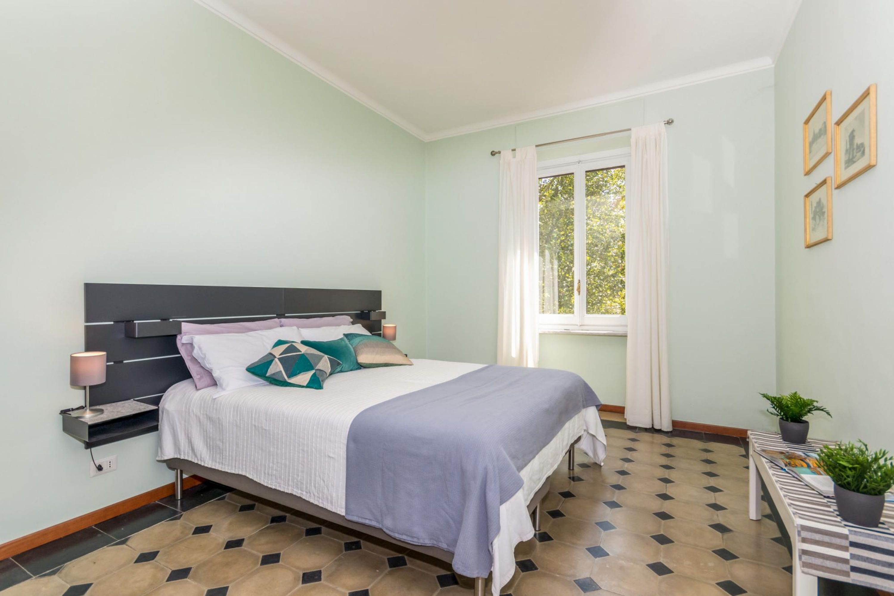 A bedroom with a bed, bedside table, lamp, window with curtains, and a view of trees.