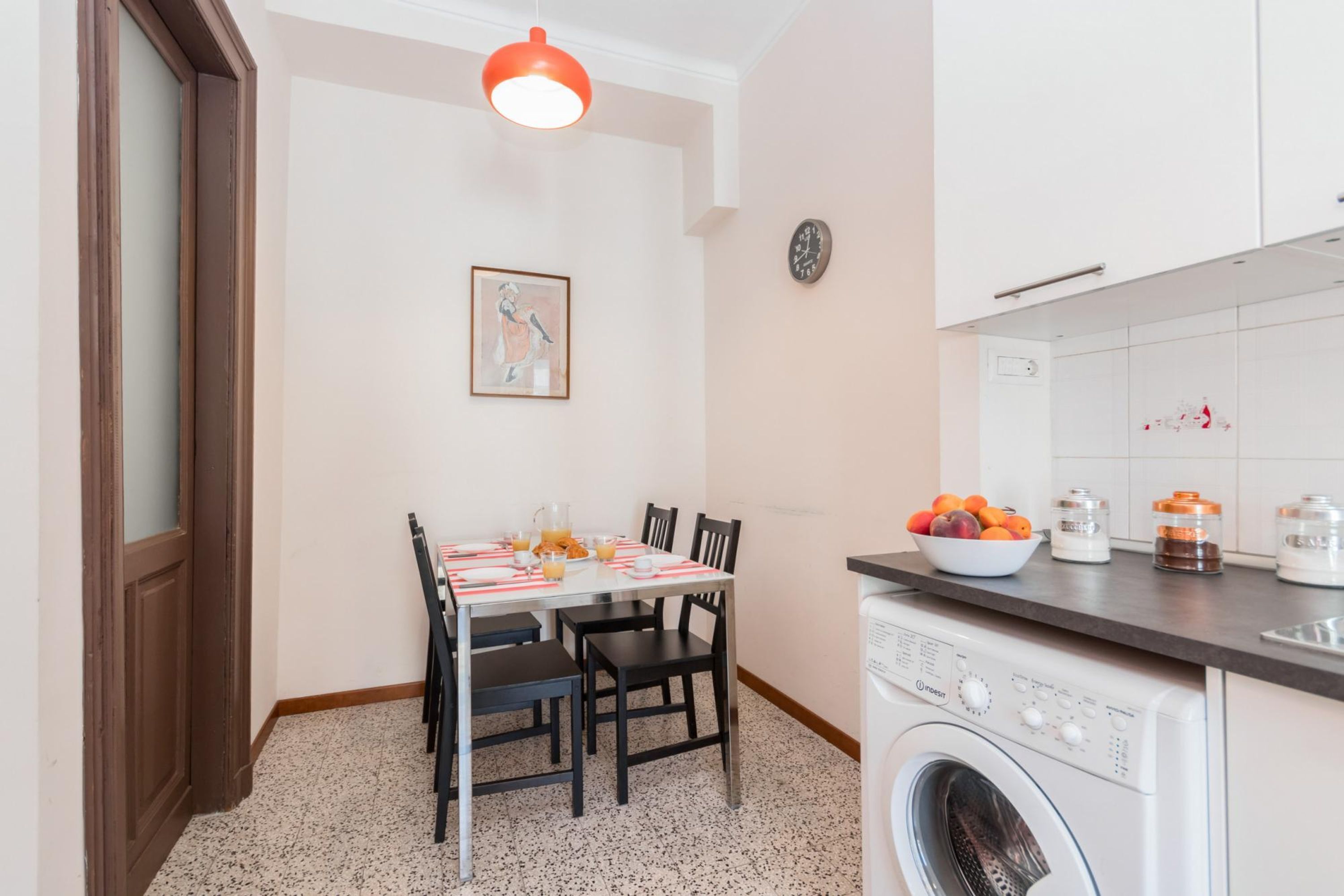 A compact kitchen with a washing machine, a dining table with chairs, and a fruit bowl on the counter.