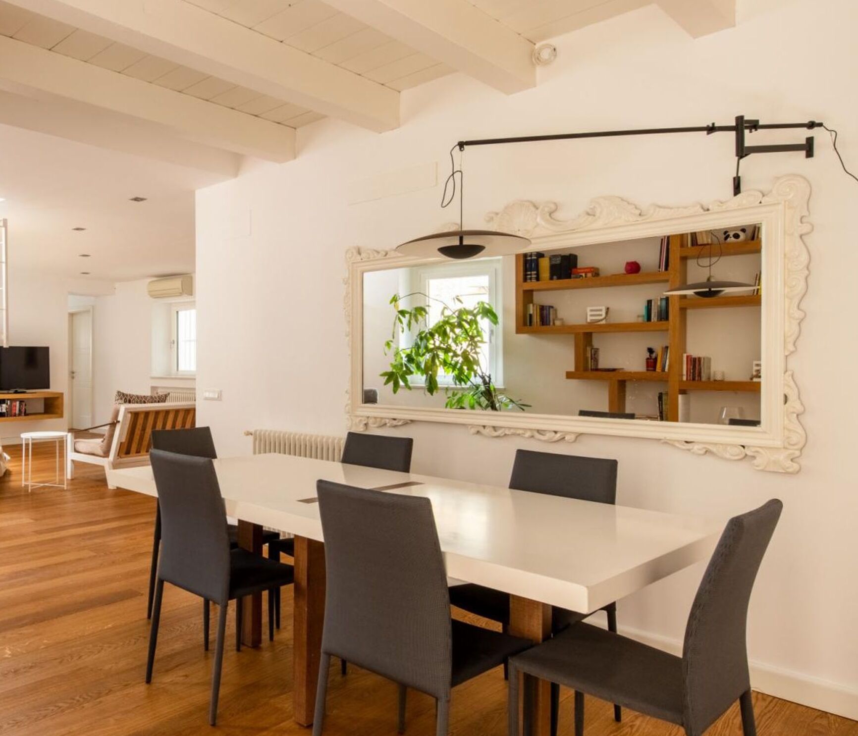 A modern dining area with a white table and black chairs, a large mirror, and a bookshelf with books and decorative items.