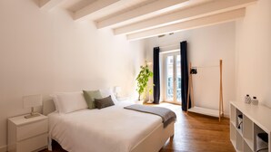 A bedroom with a large bed, bedside tables, a nightstand, a plant, a window with curtains, and a wooden floor.