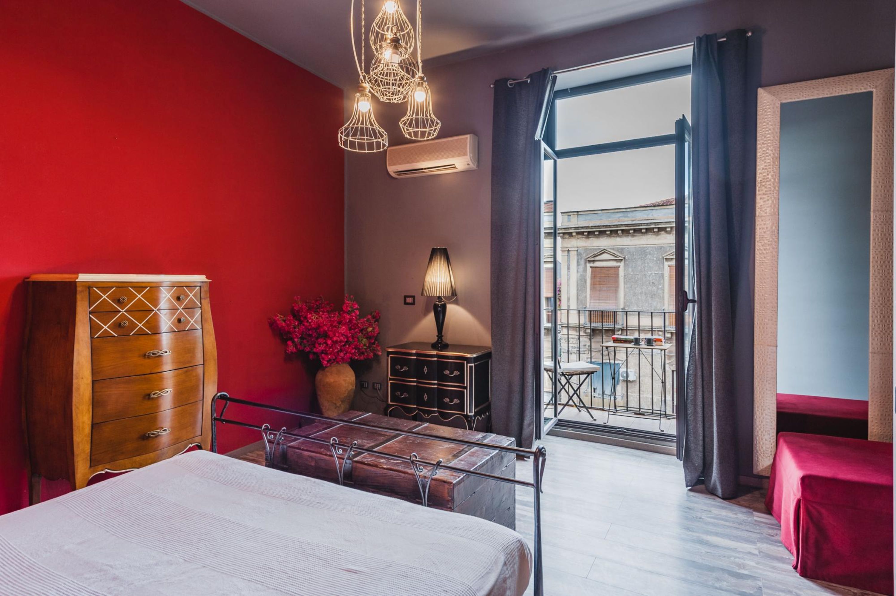 A hotel room with a bed, a dresser, a lamp, a vase with flowers, and a view of the outside.