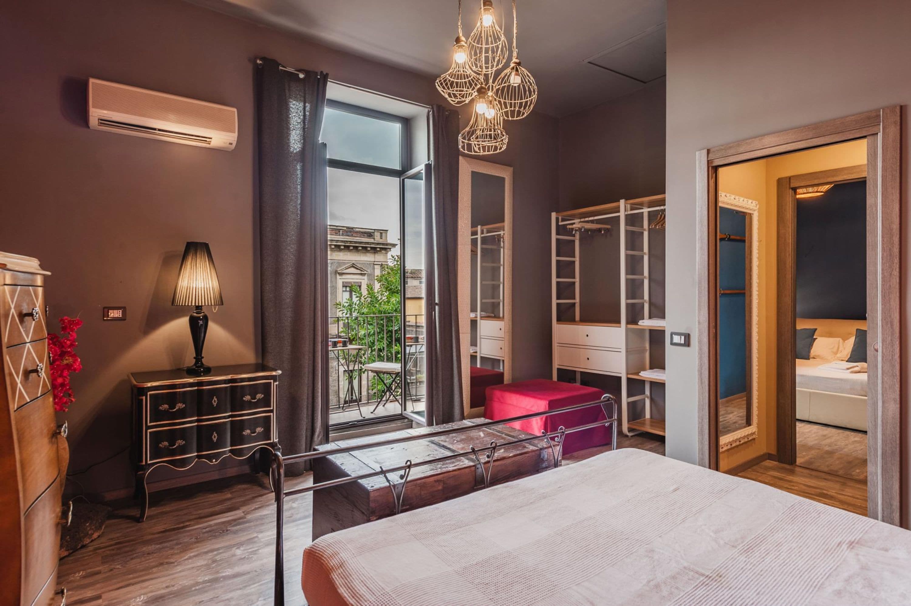 A bedroom with a bed, a dresser, a lamp, a closet, and a window with a view of buildings.