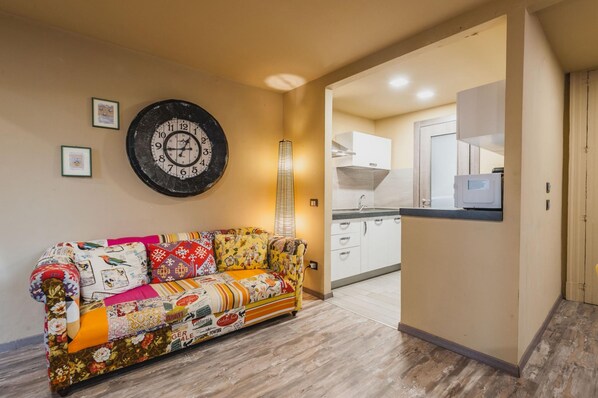 A living room with a sofa, a wall clock, and a kitchen area with cabinets and a countertop.