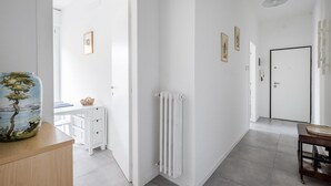 Een lichte hal met witte wanden, een radiator en een deur. Er staat een klein tafeltje met een mand en een vaas met een ontwerp van een boom.
