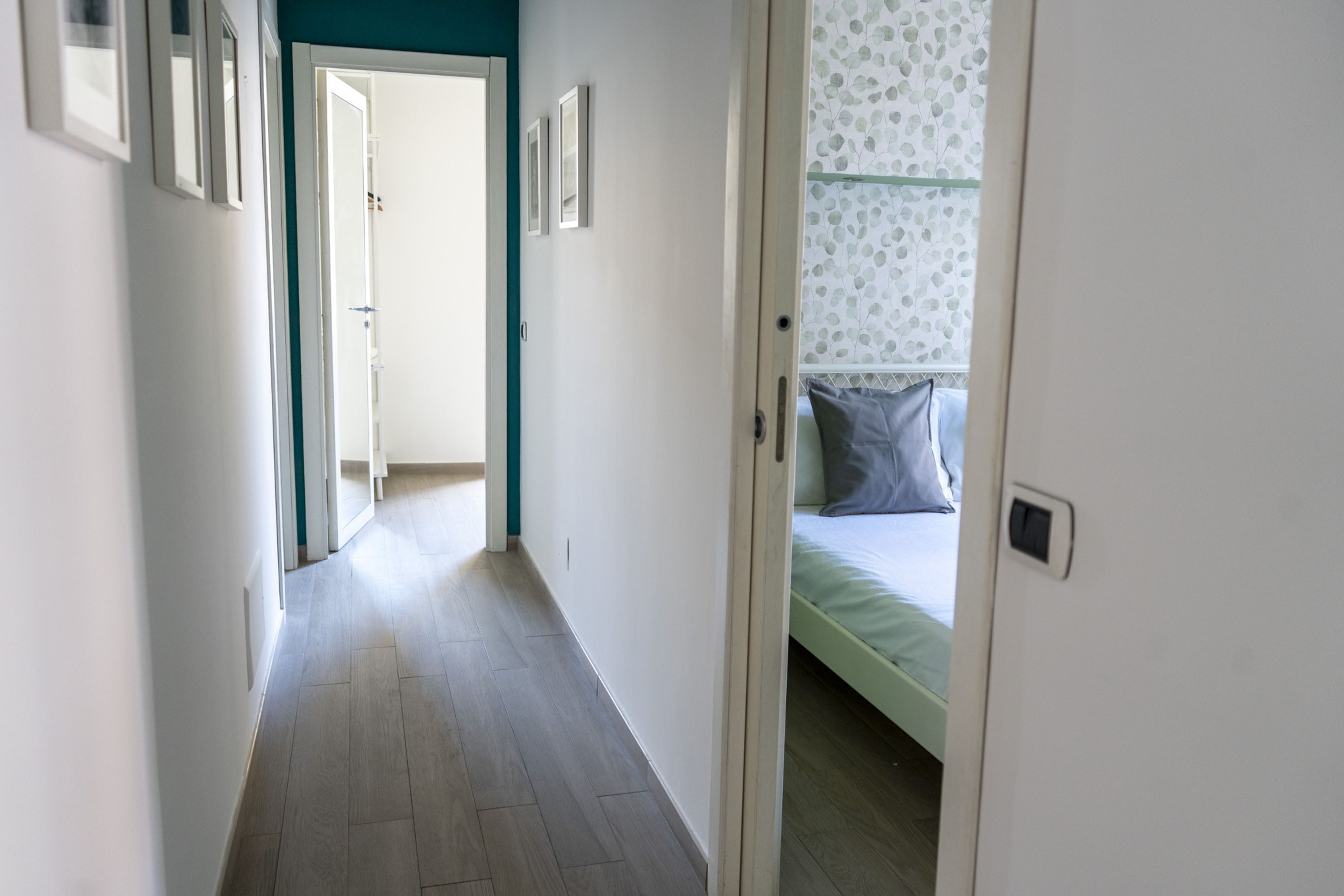 A hallway leading to a bedroom with a bed, a door, and a wall with framed pictures.