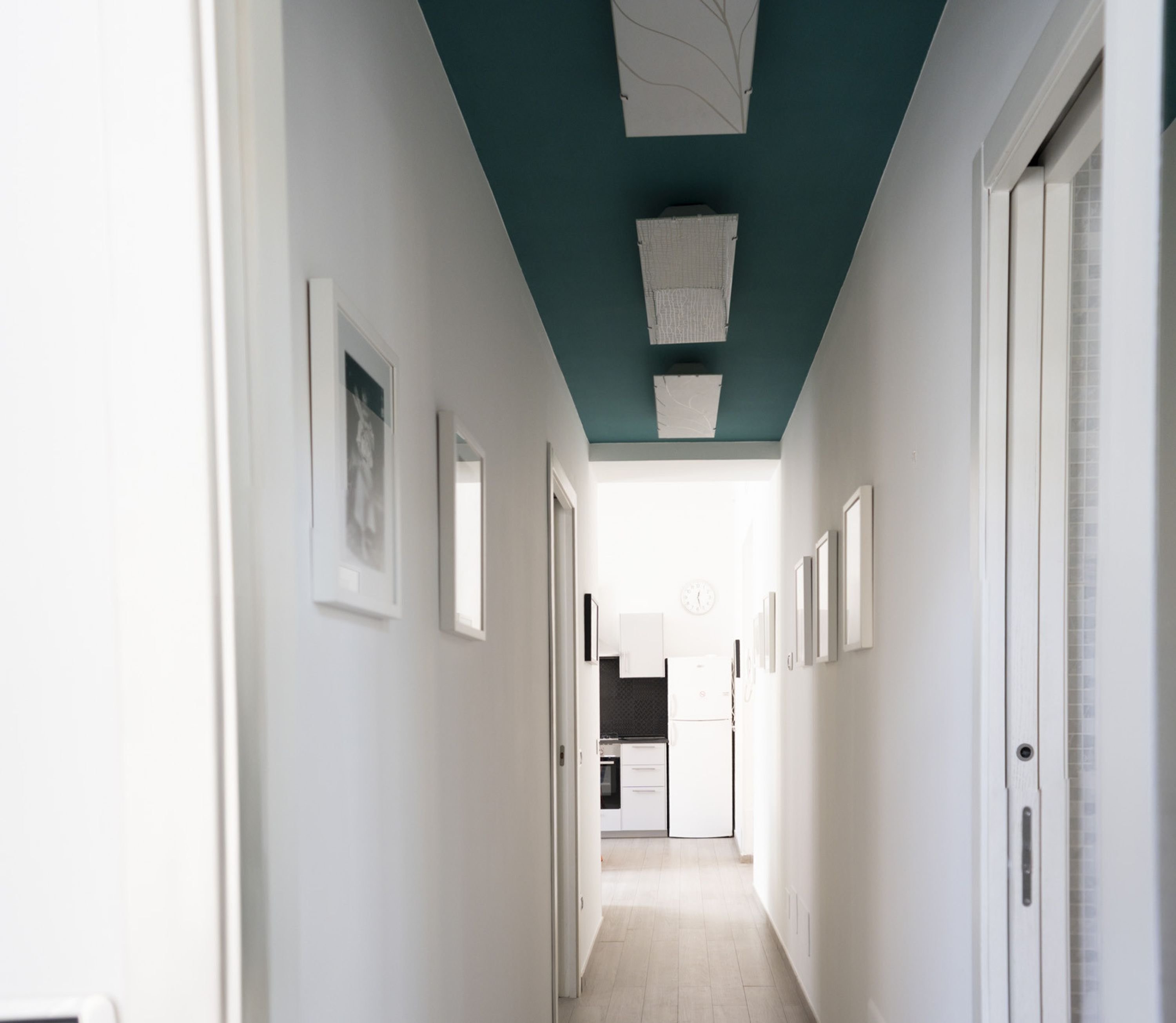 Un couloir étroit aux murs blancs, avec un plafond de couleur turquoise et une cuisine visible au fond.