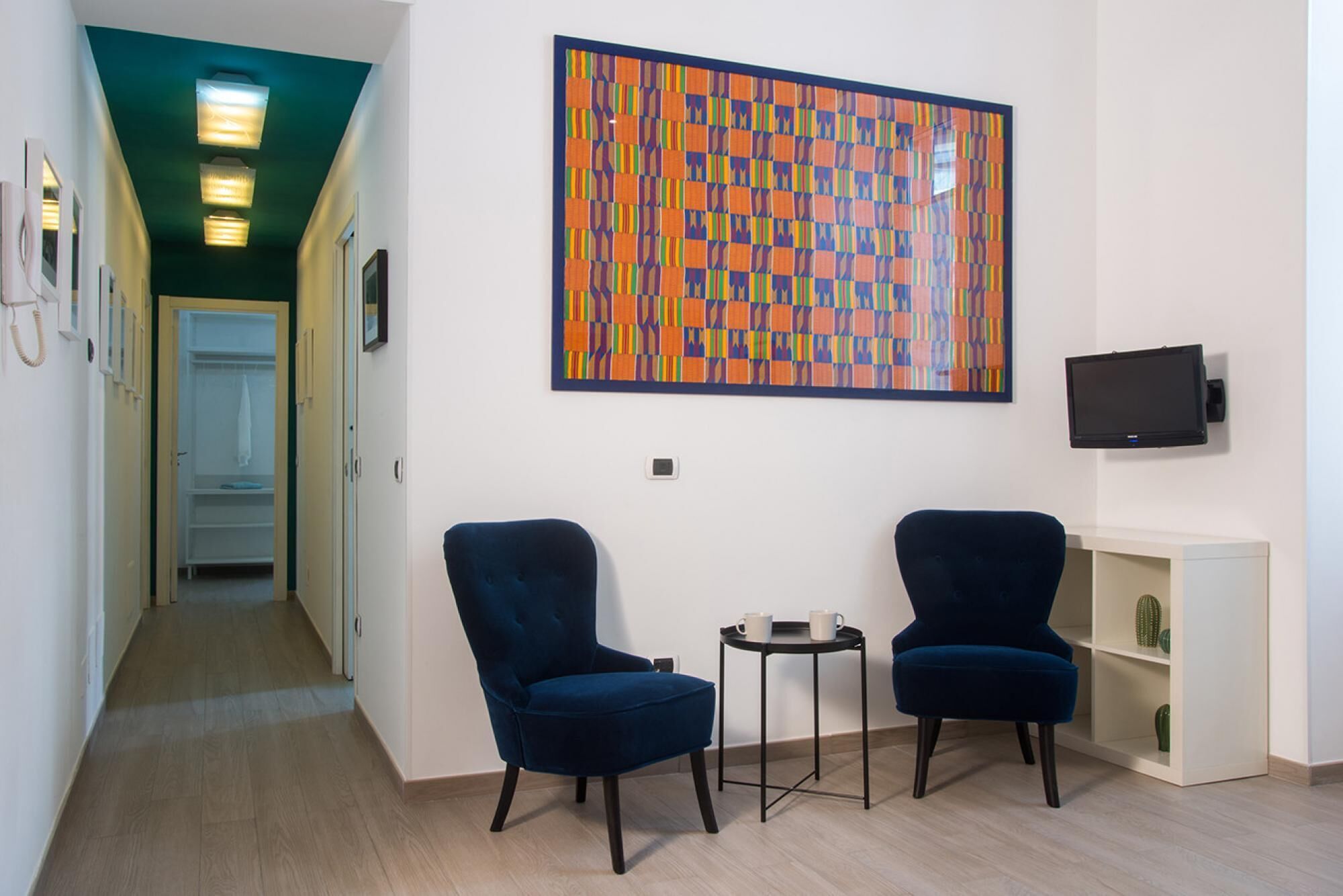 A modern room with two blue armchairs, a small table with two cups, a colorful abstract painting, a television, and a bookshelf with a cactus.