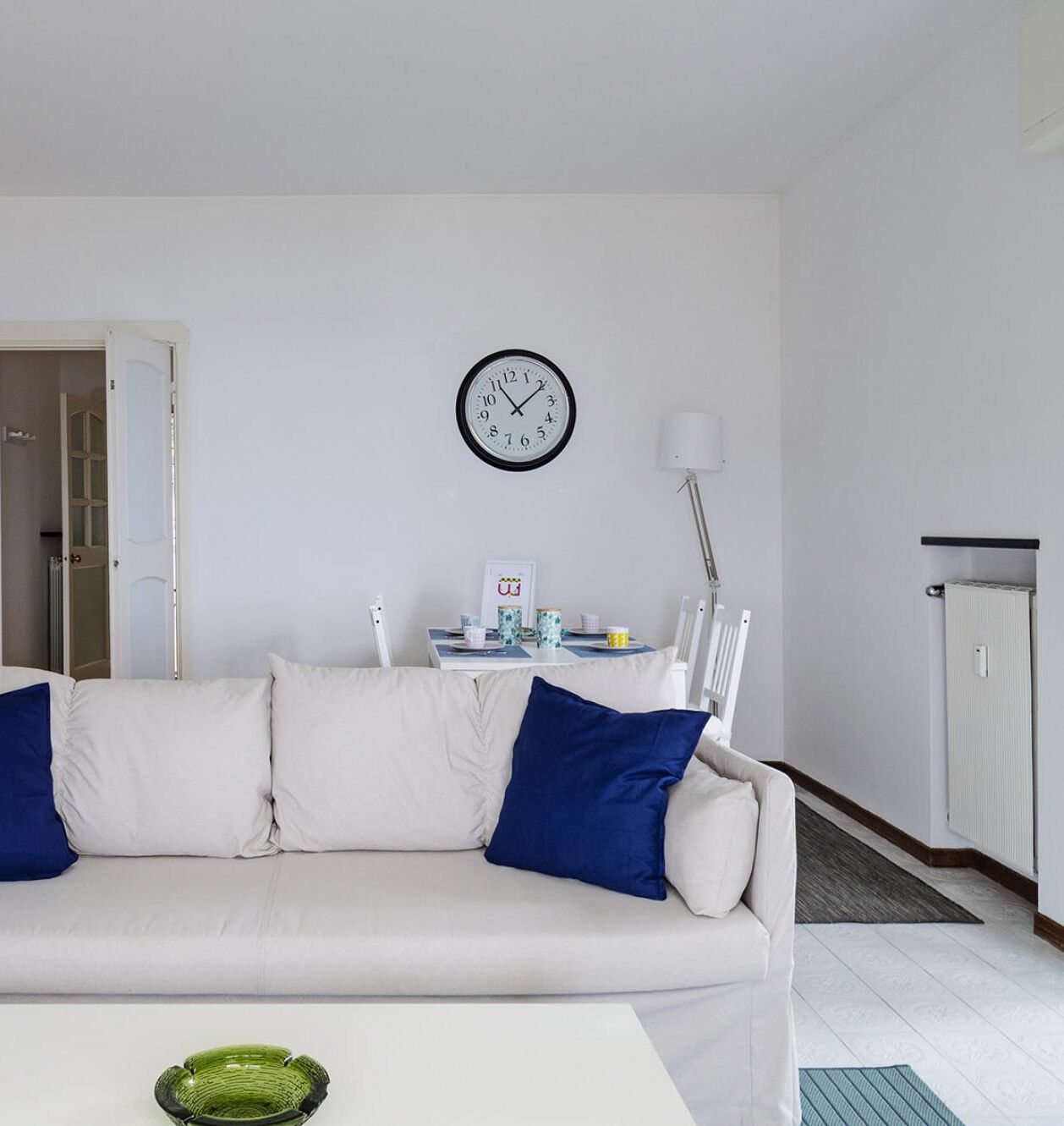 A living room with a white sofa, blue cushions, a green bowl, a clock, a table with a lamp, and a radiator.