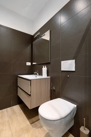 Une salle de bain moderne avec des murs recouverts de carreaux foncés, un meuble-lavabo en bois, des toilettes blanches et un miroir.