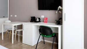 A compact hotel room with a kitchenette, a flat-screen TV mounted on the wall, a white desk, a green chair, and a potted plant.