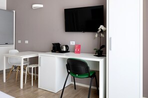 Une chambre d’hôtel compacte comprenant un coin cuisine, un téléviseur à écran plat fixé au mur, un bureau blanc, une chaise verte et une plante en pot.