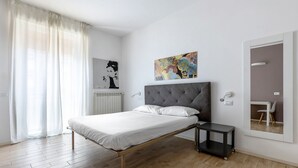 A modern bedroom with a bed, a small bedside table, a radiator, a wall-mounted artwork, and a mirror reflecting a desk and chair.